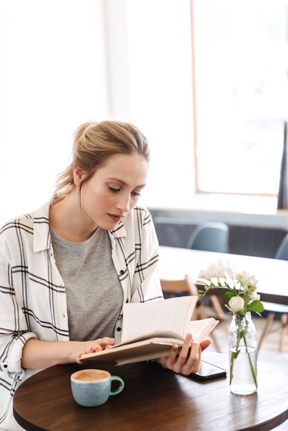 Bella giovane donna che legge il libro seduti al chiuso a bere il caffè