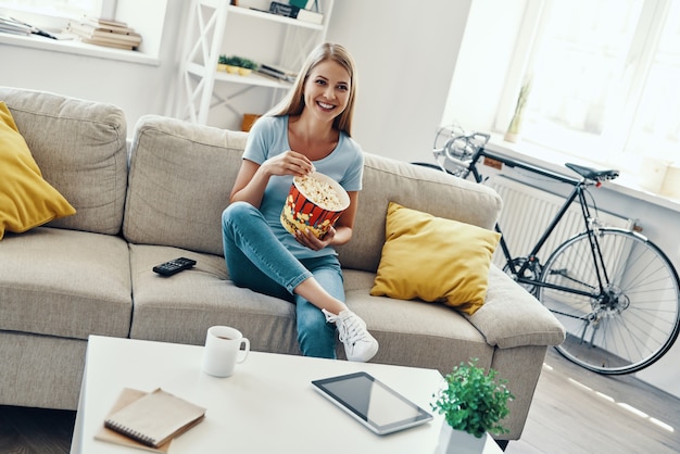 Bella giovane donna che mangia popcorn e sorride mentre guarda la TV sul divano di casa