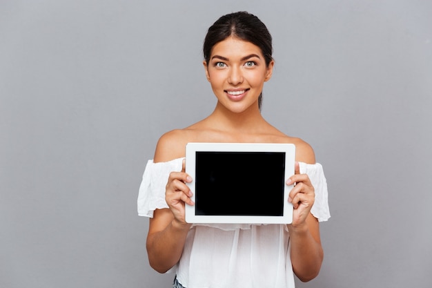 Bella giovane donna che mostra computer tablet con schermo vuoto isolato su un muro grigio