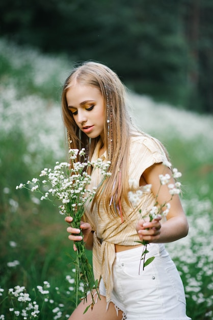 Bella giovane donna che raccoglie fiori di campo bianchi sullo sfondo di un paesaggio forestale in estate