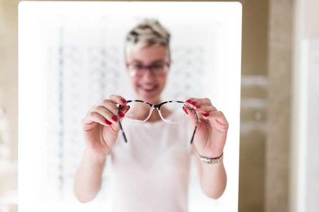 Bella giovane donna che sceglie il telaio degli occhiali nel negozio di ottica. Messa a fuoco selettiva sulle mani.