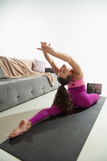 Bella giovane donna che si allena al chiuso facendo esercizio di yoga sul tappetino grigio a casa