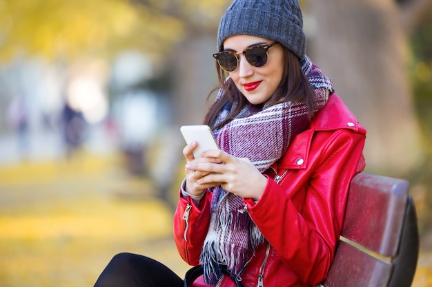 Bella giovane donna che si siede in una panchina e usando il suo telefono cellulare in autunno.