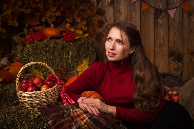 Bella giovane donna che si siede nella regolazione di autunno sul fieno con un cestino delle mele e della sorba