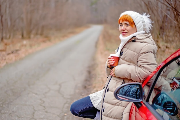 Bella giovane donna che sorride, bevendo caffè da una tazza di carta accanto alla macchina rossa sullo sfondo della foresta di autunno