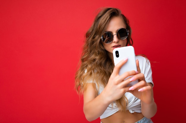 Bella giovane donna che tiene il telefono cellulare prendendo selfie foto utilizzando la fotocamera dello smartphone indossando