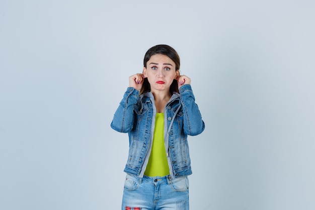 Bella giovane donna che tira giù i lobi delle orecchie, curva il labbro inferiore in abito di jeans e sembra spaventata, vista frontale.