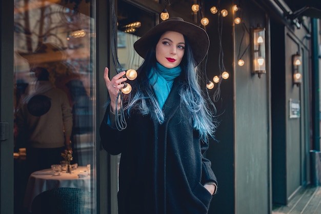 Bella giovane donna con capelli blu in bel cappotto nero, jeans e cappello.