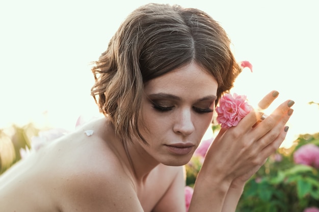 Bella giovane donna con capelli ricci che posano vicino alle rose in un giardino. Il concetto di pubblicità del profumo.