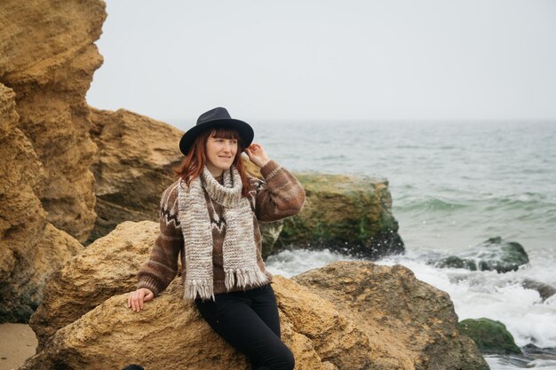 Bella giovane donna con cappello e sciarpa in piedi su una roccia contro un bellissimo mare all'orizzonte