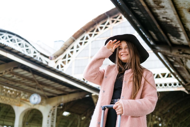 Bella giovane donna con cappello nero e cappotto rosa con valigia si trova sulla piattaforma della stazione tou...