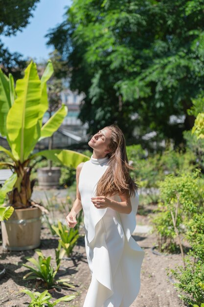 Bella giovane donna con i capelli lunghi in abito bianco che cammina all'aperto godendosi la luce del sole con gli occhi chiusi Giardino tropicale estivo