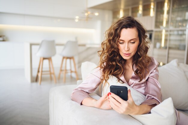 Bella giovane donna con i capelli ricci in pigiama rosa a divano bianco al mattino. Signora che utilizza smartphone nell'interno del salone di stile scandinavo.