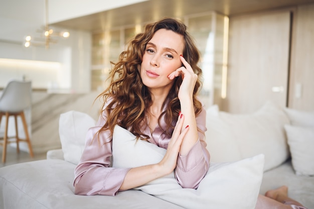 Bella giovane donna con i capelli ricci in pigiama rosa seduto al divano bianco o divano al mattino. Soggiorno in stile scandinavo e interni cucina.