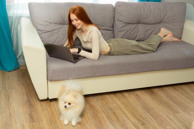 bella giovane donna con i capelli rossi si trova a casa sul divano con un computer portatile