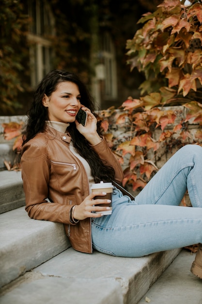 Bella giovane donna con il cellulare beve caffè per andare alle scale all'aperto il giorno d'autunno