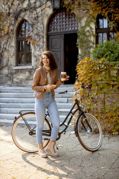 Bella giovane donna con il cellulare beve caffè per andare in bicicletta il giorno d'autunno