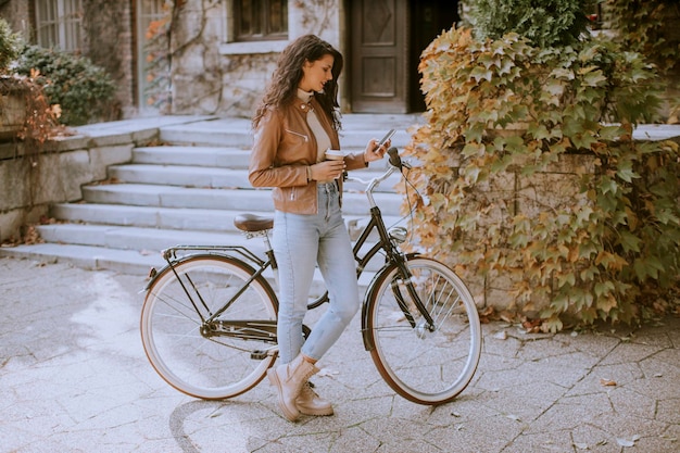 Bella giovane donna con il cellulare beve caffè per andare in bicicletta il giorno d'autunno