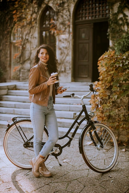 Bella giovane donna con il cellulare beve caffè per andare in bicicletta il giorno d'autunno