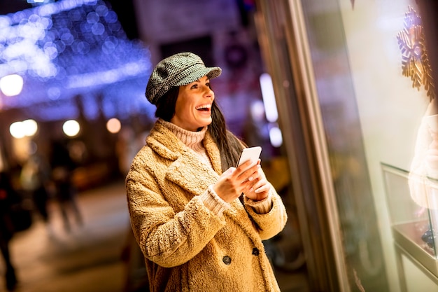 Bella giovane donna con il cellulare vicino alla vetrina del negozio a Natale