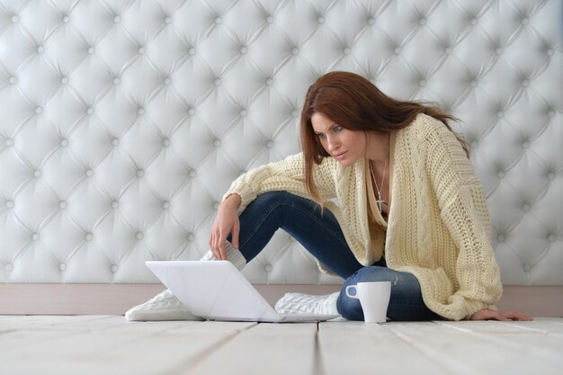 Bella giovane donna con il computer portatile seduto sul pavimento e bere caffè