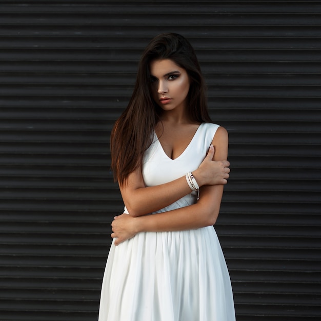 Bella giovane donna con il trucco in vestito bianco vicino alla parete scura del metallo