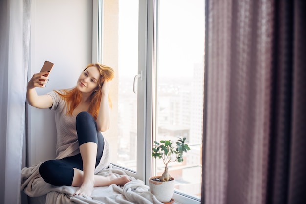 Bella giovane donna con lunghi capelli rossi si fa un selfie sul suo smartphone mentre è seduta sul davanzale. Bella ragazza con un telefono in un ambiente domestico. Concetto di tecnologia e social network.