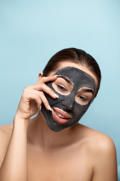 Bella giovane donna con maschera per il viso di argilla nera su pelle pulita fresca Ragazza modello bellezza cura del viso con maschera cosmetica idratante tocca il viso Trattamento viso Cura della pelle