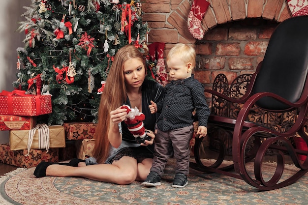 Bella giovane donna con un bambino di un anno che gioca vicino all'albero di Natale nel soggiorno. Mamma con figlio carino nella stanza decorata di Natale. Sono sorridenti e felici. Concetto di Capodanno in famiglia. Copia spazio