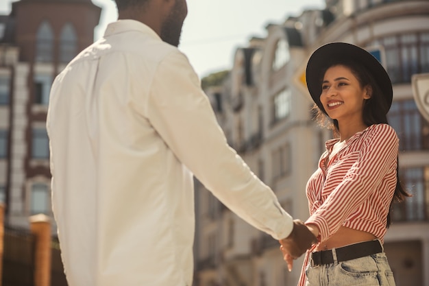 Bella giovane donna con un cappello nero che sorride al suo ragazzo e gli tiene la mano all'aperto