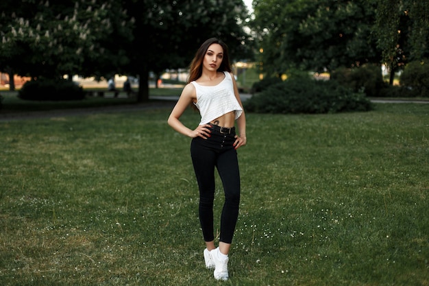 Bella giovane donna con un corpo in una maglietta bianca, jeans neri e scarpe bianche in posa nel parco