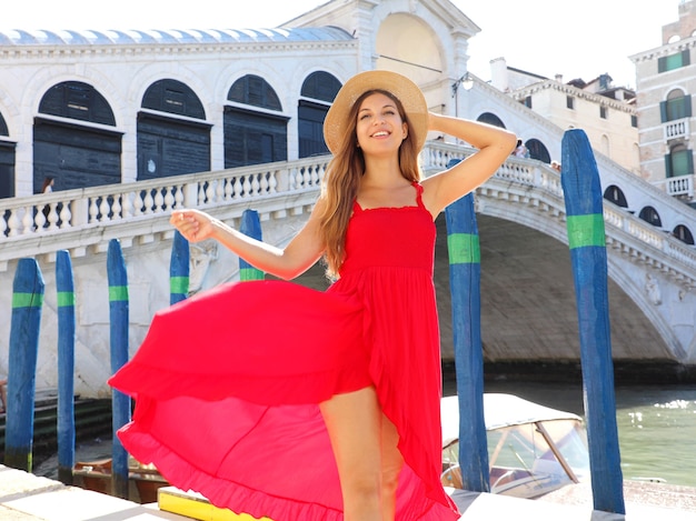 Bella giovane donna con un vestito rosso è in piedi davanti al famoso Ponte di Rialto a Venezia, Italia