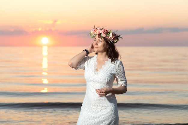 Bella giovane donna con una corona di fiori sulla testa in riva al mare al tramonto