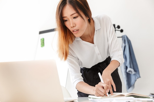 Bella giovane donna d'affari asiatica in piedi alla sua scrivania in ufficio, lavorando su un computer portatile