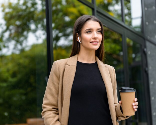 Bella giovane donna d'affari che indossa cappotto autunnale che cammina all'aperto, tenendo una tazza di caffè da asporto