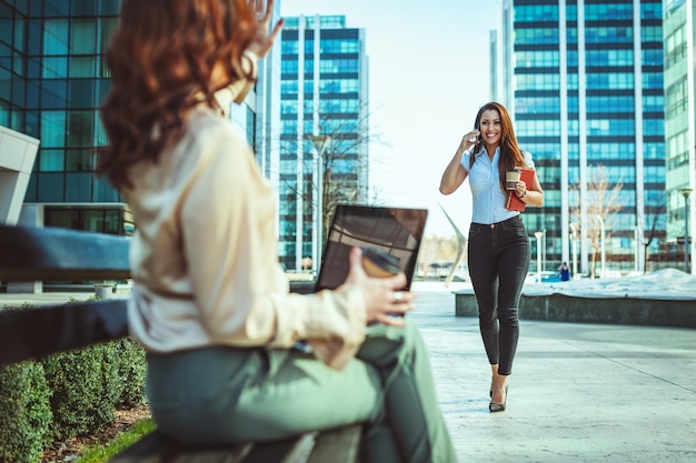 Bella giovane donna d'affari è seduta sulla panchina e tiene in mano il laptop sullo sfondo della città urbana e il suo collega, che sta venendo da lei, sta parlando con lo smartphone.