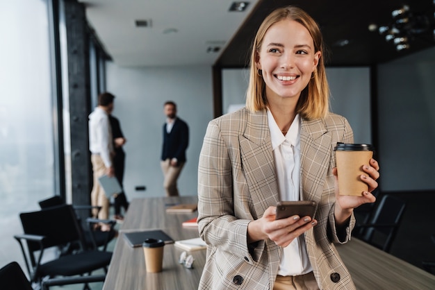 Bella giovane donna d'affari intelligente sorridente in piedi in ufficio con un gruppo di colleghi sul muro, usando il telefono cellulare, tenendo una tazza di caffè da asporto