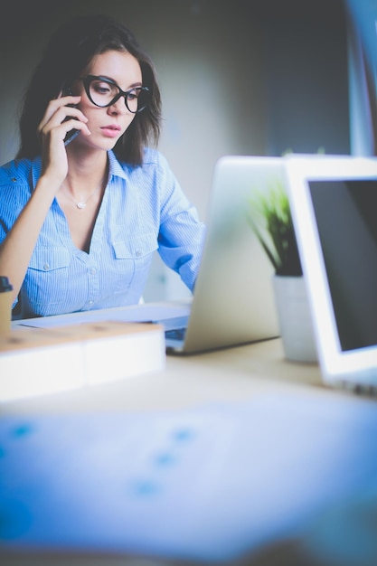 Bella giovane donna d'affari seduto alla scrivania in ufficio e parlando al cellulare Donna d'affari