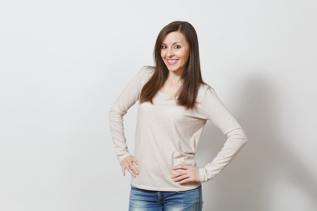 Bella giovane donna dai capelli castani felice europea con pelle sana e pulita, vestita con abiti casual leggeri, jeans che guarda l'obbiettivo con timido sorriso affascinante su sfondo bianco. Concetto di emozioni.