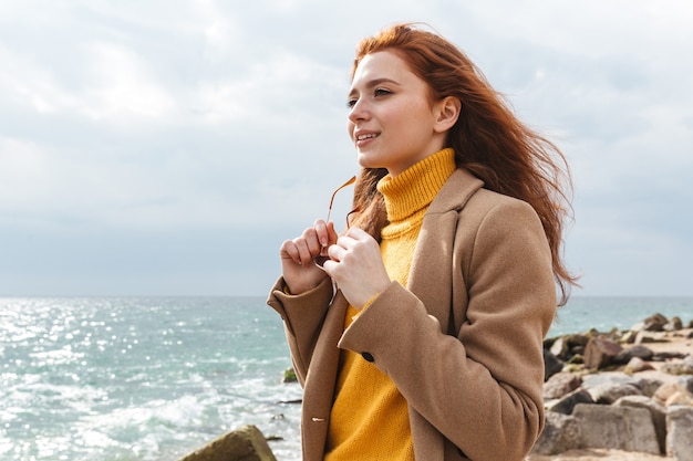 Bella giovane donna dai capelli rossi che indossa un cappotto che cammina sulla spiaggia