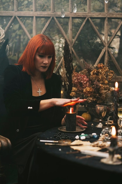 Bella giovane donna dai capelli rossi con cappello nero e vestito da strega fa magie in serra decorata per la celebrazione di Halloween con zucche e teschio nel fumo
