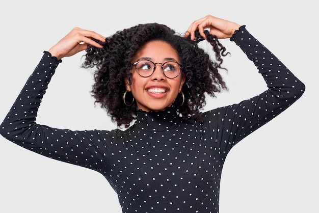 Bella giovane donna dalla carnagione scura vestita di nero con camicetta a pois bianchi che si tiene per mano sui capelli sentendosi felice femmina afroamericana sorridente largamente indossando occhiali rotondi in posa in studio