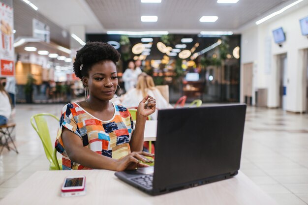 Bella giovane donna dalle free lance dalla pelle scura che per mezzo del computer portatile che si siede alla Tabella del caffè. Concetto di lavoro freelance.