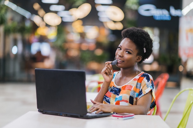 Bella giovane donna dalle free lance dalla pelle scura che per mezzo del computer portatile che si siede alla Tabella del caffè. Concetto di lavoro freelance.