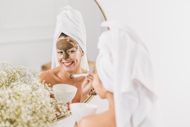 Bella giovane donna davanti allo specchio che applica la maschera facciale al salone di bellezza.