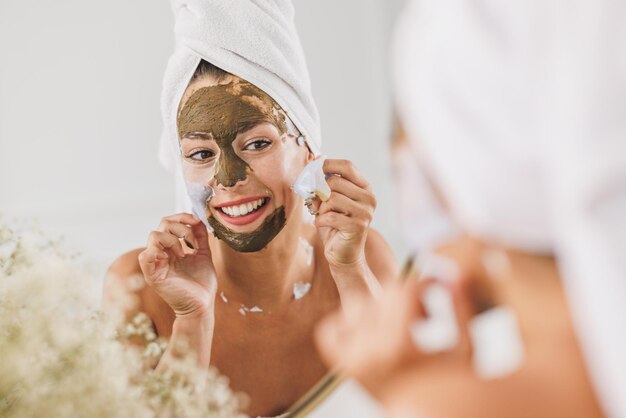 Bella giovane donna davanti allo specchio che rimuove la maschera facciale nel bagno di casa.