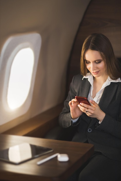 Bella giovane donna di affari che viaggia in jet privato. Signora dai capelli castani in camicetta bianca, abito formale con smartphone, seduta in un comodo sedile in aereo.