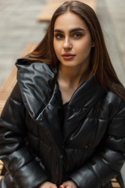 Bella giovane donna di moda in un elegante piumino invernale nero si siede su una panchina e guarda la telecamera