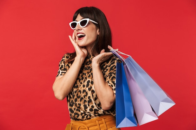 bella giovane donna eccitata vestita con una camicia stampata animale in posa isolata sul muro rosso indossando occhiali da sole che tengono le borse della spesa.