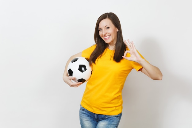 Bella giovane donna europea, tifoso o giocatore in uniforme gialla che tiene il pallone da calcio che mostra gesto OK isolato su sfondo bianco. Sport, giocare a calcio, salute, concetto di stile di vita sano.